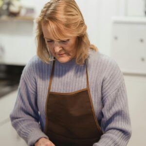 Concentrated middle aged lady in casual clothes and apron standing near counter while using knife to cut tomato on cutting board in light kitchen near metal bowl