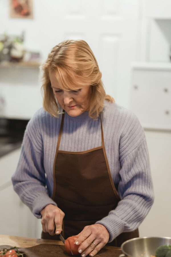 Concentrated middle aged lady in casual clothes and apron standing near counter while using knife to cut tomato on cutting board in light kitchen near metal bowl