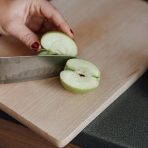 From above of crop faceless female cutting fresh juicy apple with sharp knife on wooden chopping board in kitchen