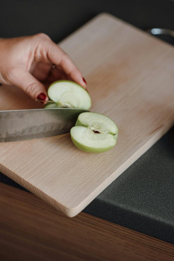 From above of crop faceless female cutting fresh juicy apple with sharp knife on wooden chopping board in kitchen